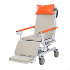 Horizontal electric patient transfer chair shown empty at a three-quarter angle with orange head pillow and armrests, padded leg rest extended and four locking casters, on a white background